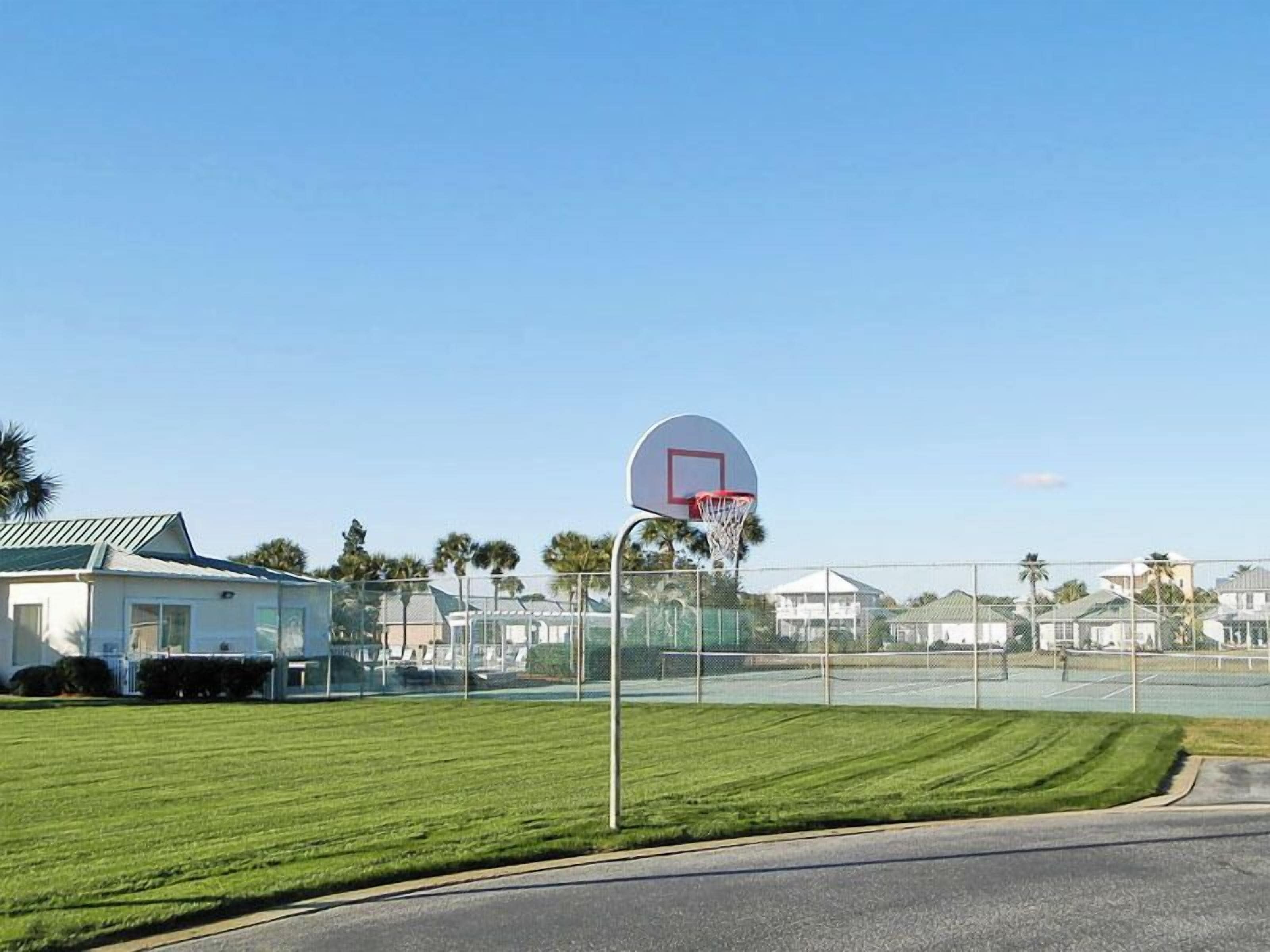 Una cancha de baloncesto con un aro, rodeada por una valla, y casas al fondo.