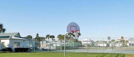 Una cancha de baloncesto con un aro, rodeada por una valla, y casas al fondo.