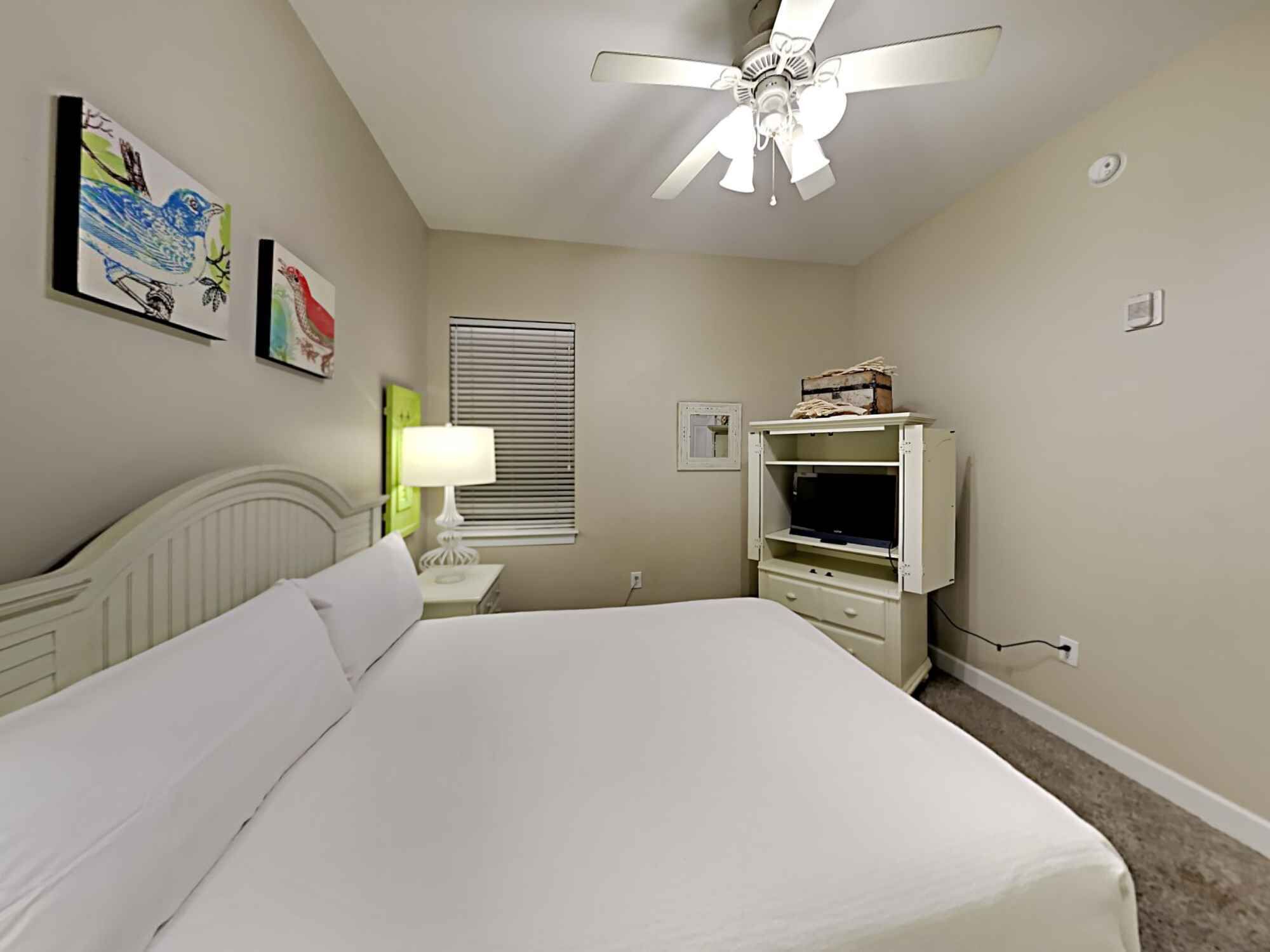 A bedroom with a bed, a ceiling fan, a nightstand with a lamp, a dresser, and wall art.