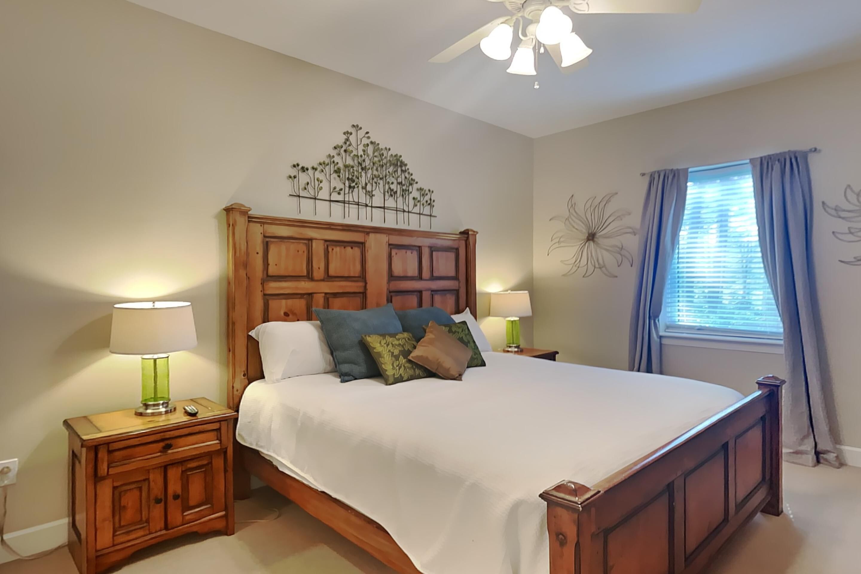 Un dormitorio con cama de madera, dos mesitas de noche con lámparas, un ventilador de techo, una ventana con cortinas y decoración en la pared.