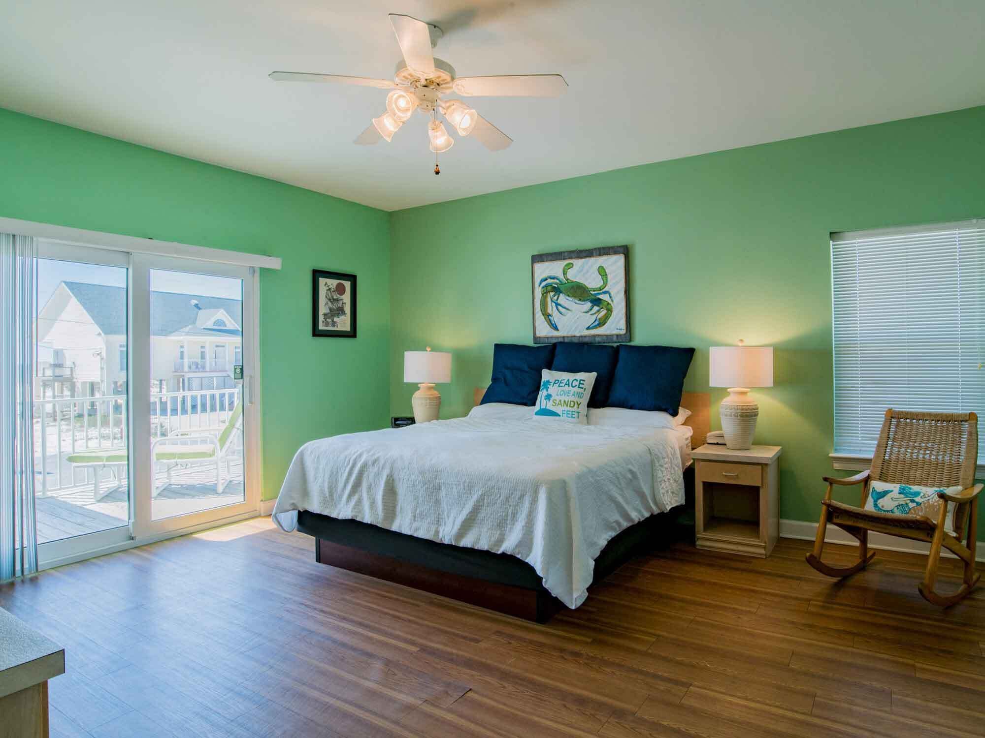 Une chambre avec un lit, deux tables de chevet, une chaise, un ventilateur de plafond et une vue sur une maison, visible à travers une porte-fenêtre coulissante.