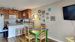 A kitchen with wooden cabinets, a microwave, and a dining area with a green table and wooden chairs. - Grand Caribbean #210 East by Southern Vacation Rentals (Destin)