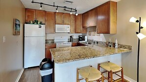A kitchen with wooden cabinets, a white refrigerator, a microwave, a stove, a sink, granite countertops, and two bar stools. - Grand Caribbean #210 East by Southern Vacation Rentals (Destin)
