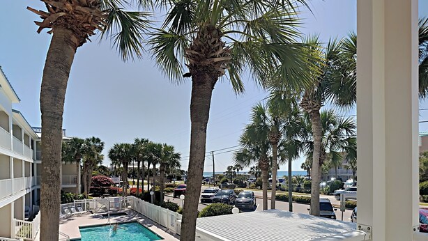 A pool area with palm trees, a pool, and a view of the street and ocean. - Grand Caribbean #210 East by Southern Vacation Rentals (Destin)