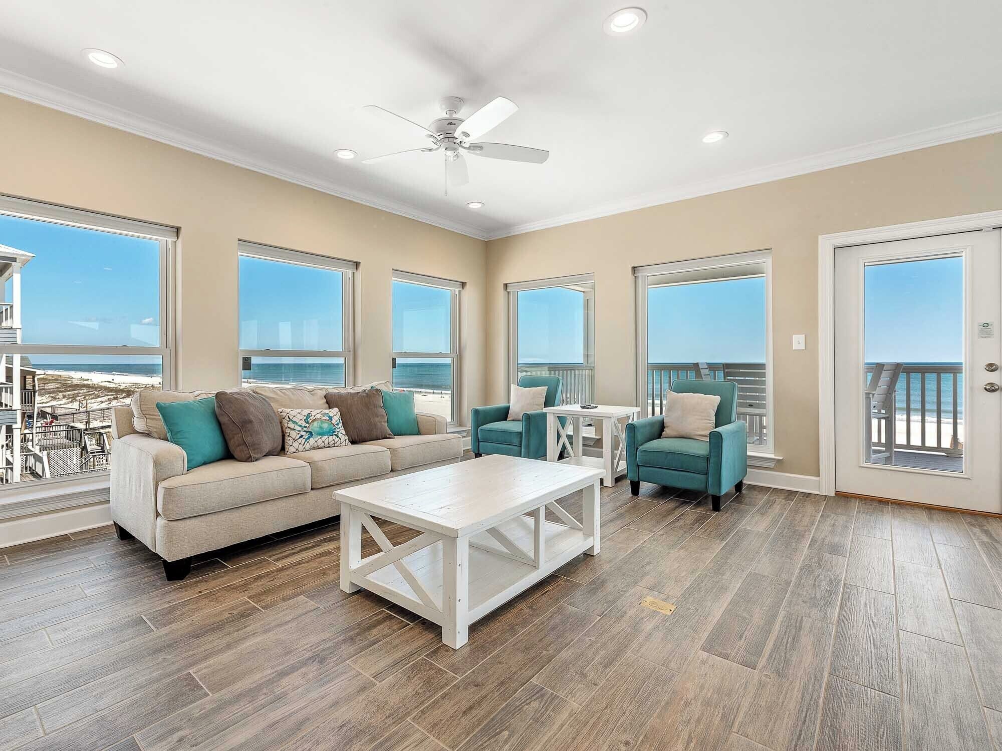 A spacious living room with a large window overlooking the beach, a ceiling fan, and a wooden floor.