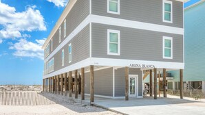 A multi-story building with a balcony area, labeled "ARENA BLANCA," featuring a coastal setting with a sandy beach and wooden posts supporting the structure. - Arena Blanca by Southern Vacation Rentals (Gulf Shores)