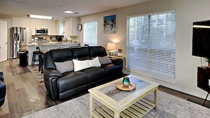 A modern living room with a black leather sofa, a wooden coffee table, and a kitchen in the background. - Dune Our Thing by Southern Vacation Rentals (Santa Rosa Beach)