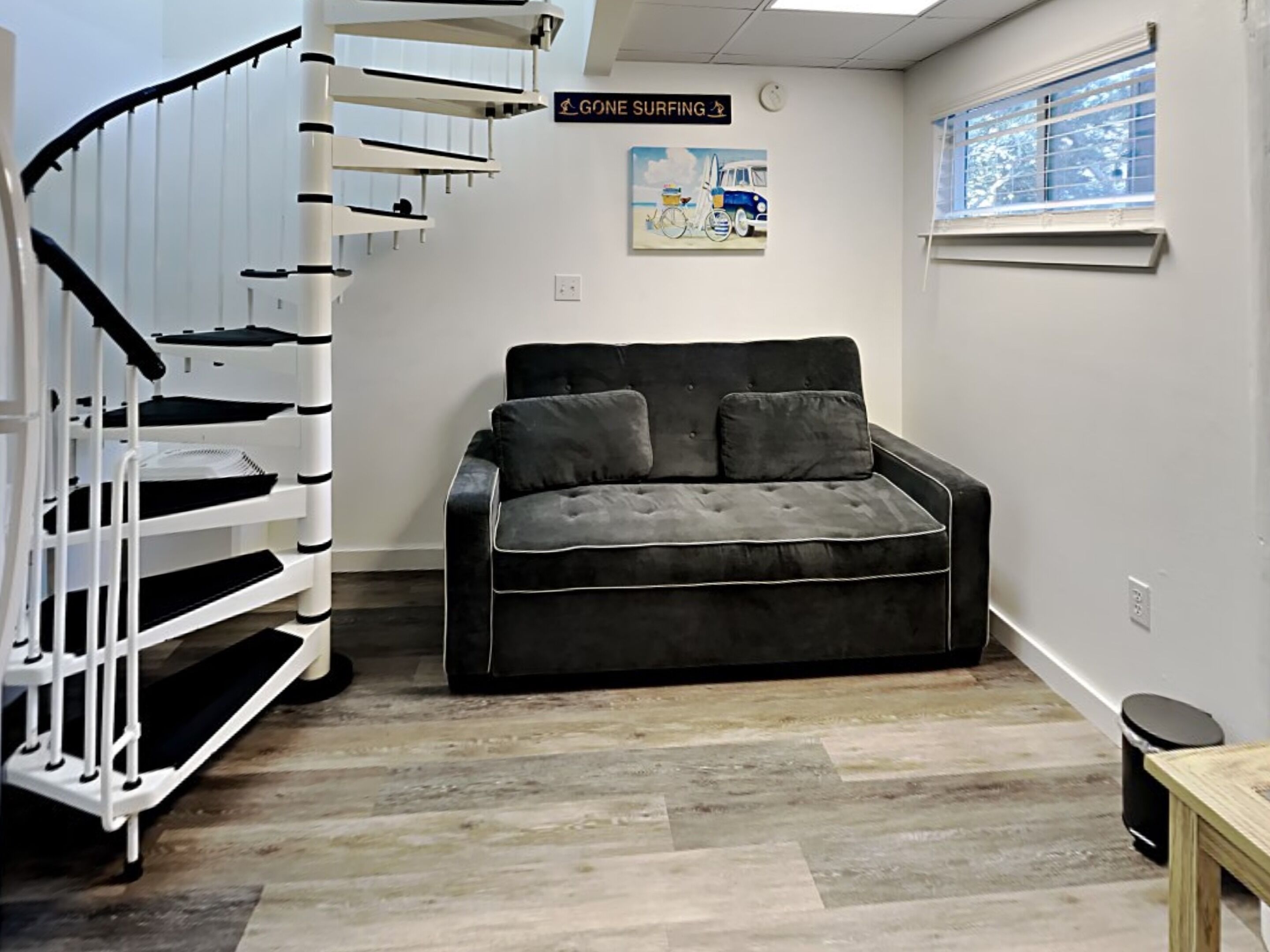 Une pièce avec un canapé noir, un escalier en colimaçon blanc, un panneau indiquant « Je pars faire du surf » et un tableau sur le thème de la plage.