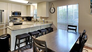 A modern kitchen with white cabinets, a granite countertop, stainless steel appliances, and a dining area with a large table and bar stools. - Dune Our Thing by Southern Vacation Rentals (Santa Rosa Beach)