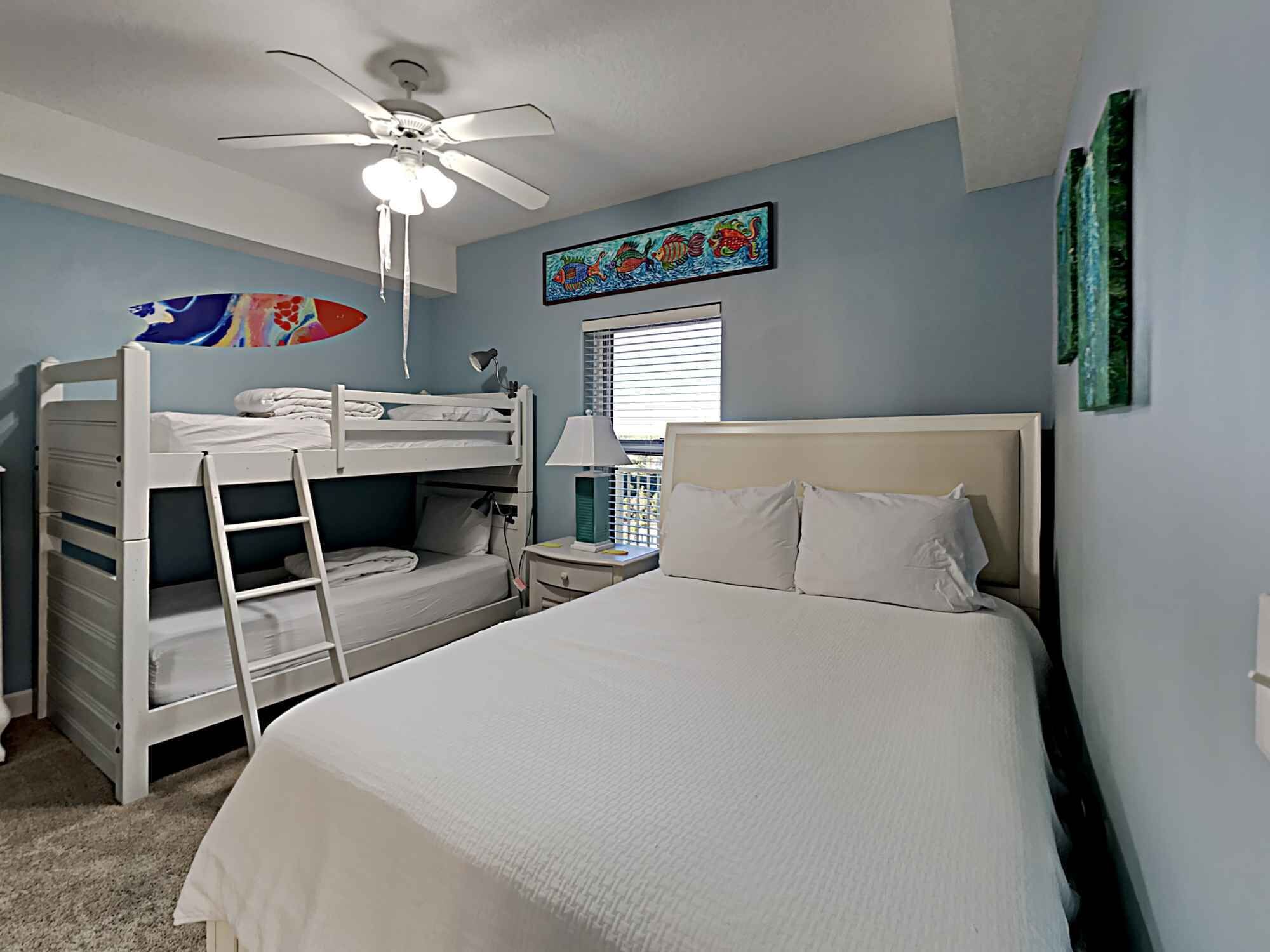 A bedroom with bunk beds, a single bed, a ceiling fan, a window with blinds, and a colorful artwork on the wall.