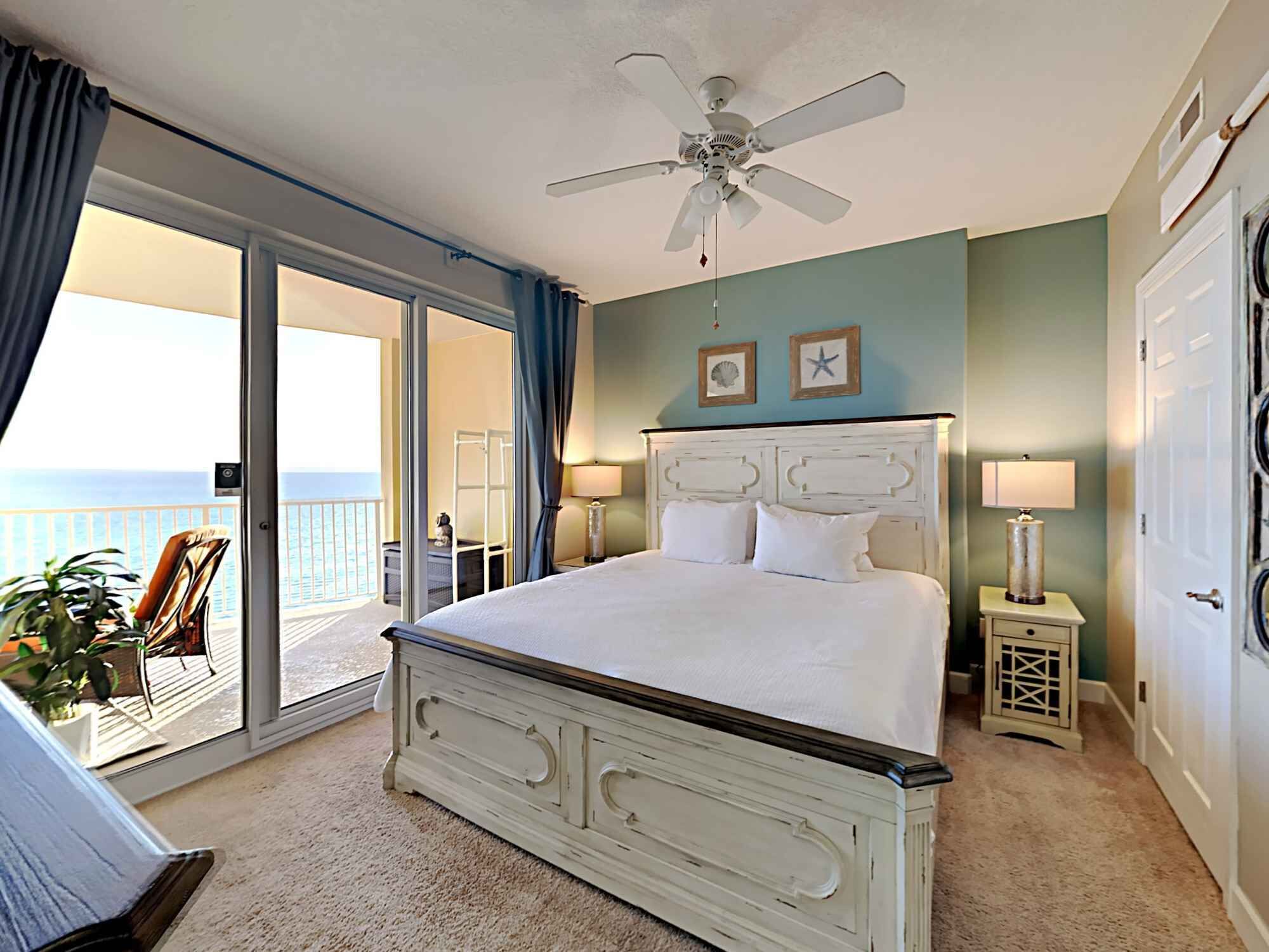 A bedroom with a large bed, a ceiling fan, a view of the ocean through a sliding door, and a small table with a lamp.