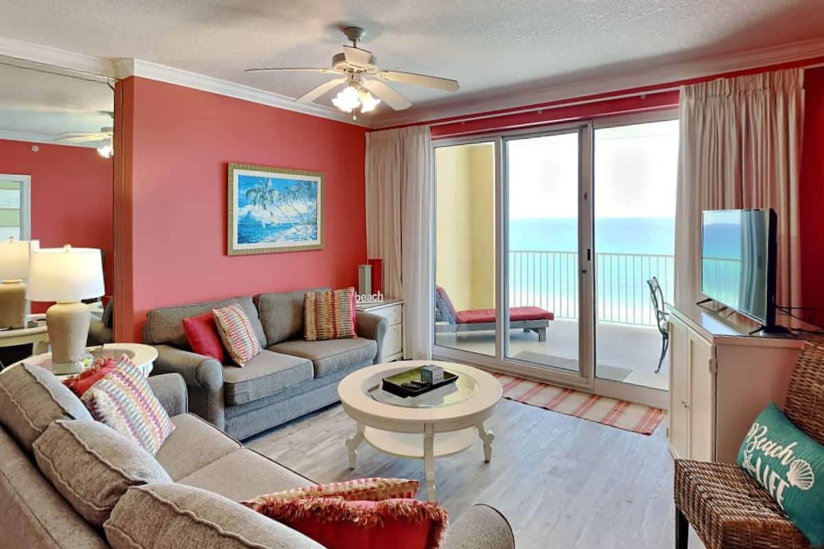 A living room with a sofa, a coffee table, and a television. There is a painting on the wall and a window with a view of the ocean.