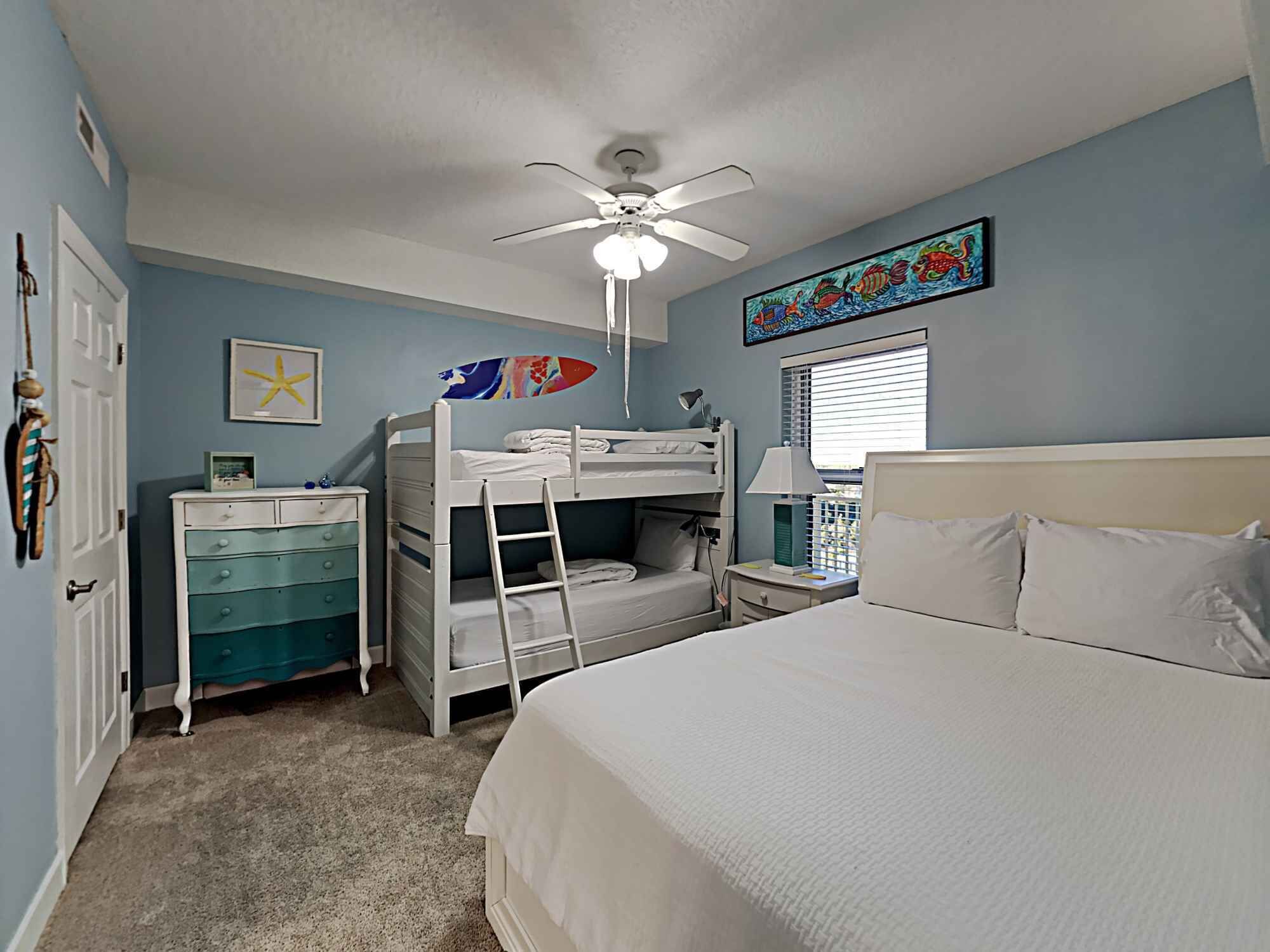 A bedroom with bunk beds, a ceiling fan, a desk, a chair, a lamp, and a window with blinds.