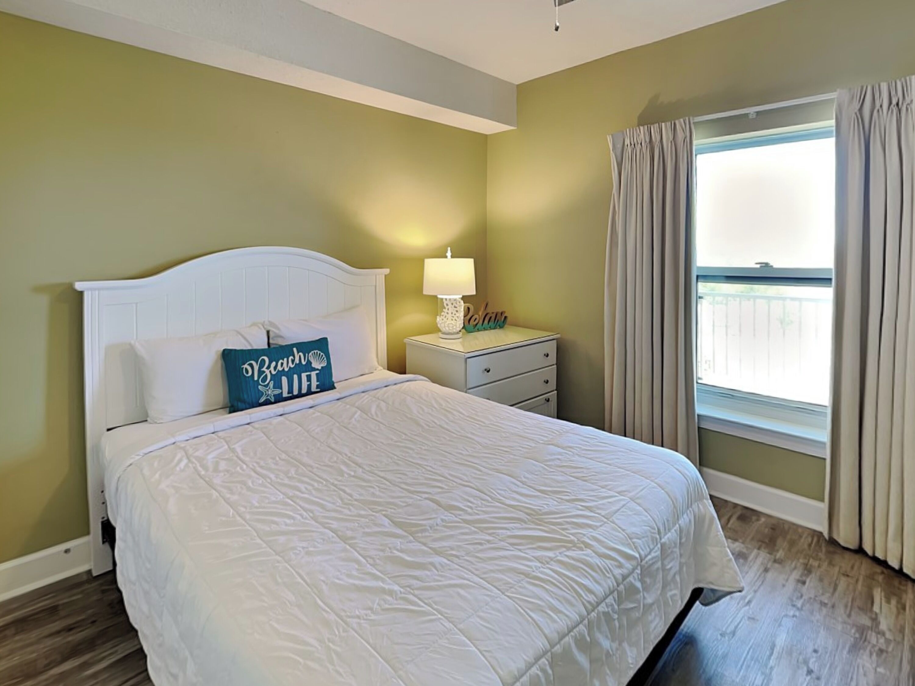 A bedroom with a white bed, a nightstand with a lamp, and a window with curtains.