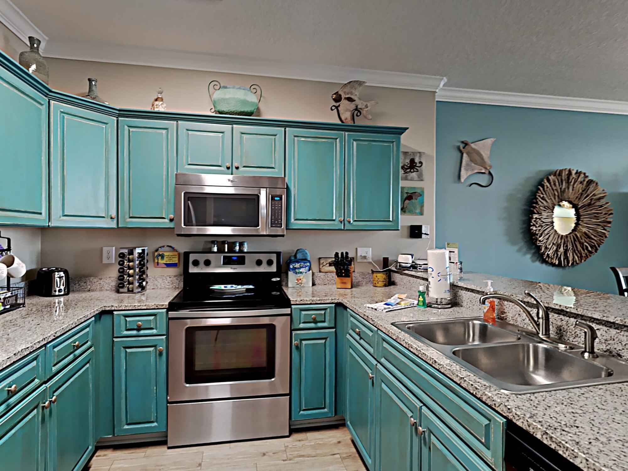 A kitchen with teal cabinets, stainless steel appliances, granite countertops, and a decorative wall piece.