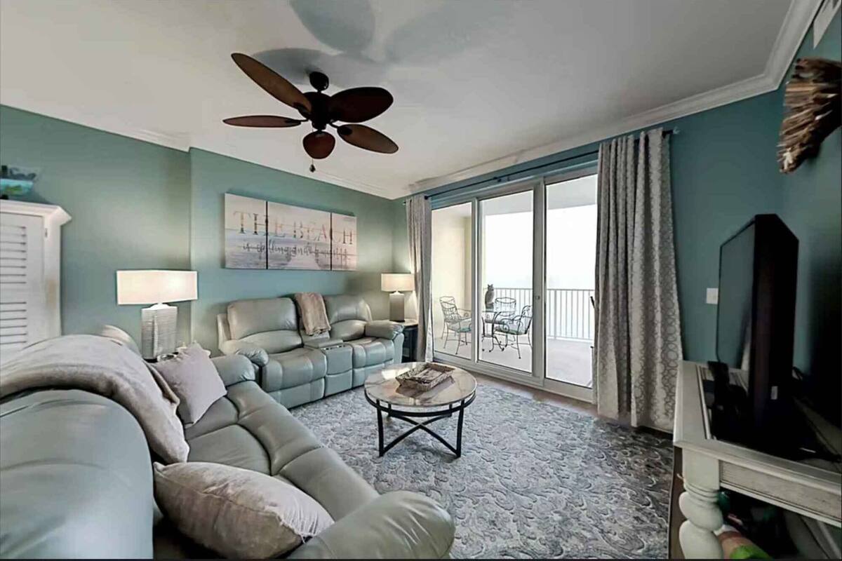 A living room with a sectional sofa, a coffee table, a ceiling fan, and a sliding glass door leading to a balcony.
