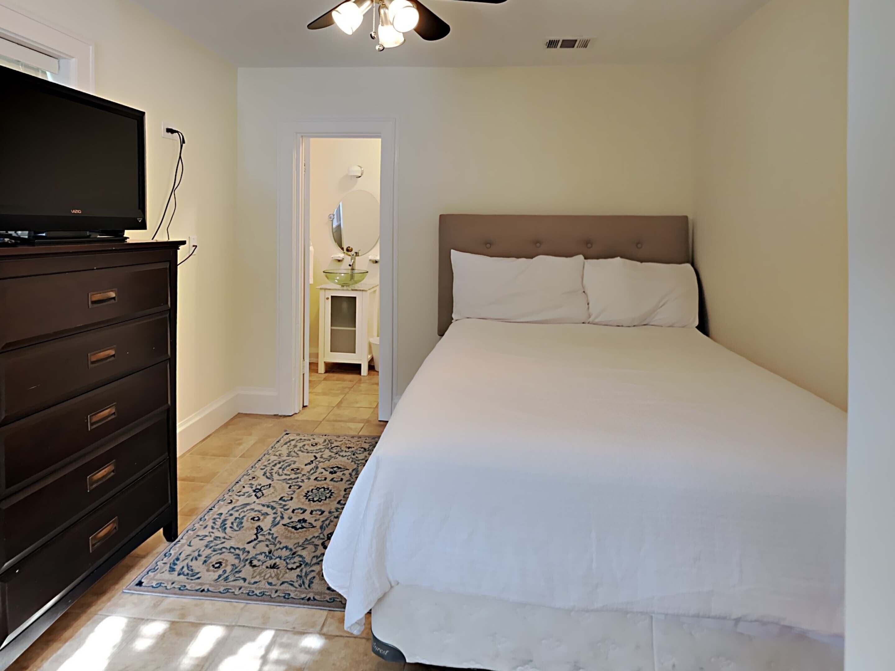 A bedroom with a bed, a dresser, a television, and a bathroom visible through an open door.