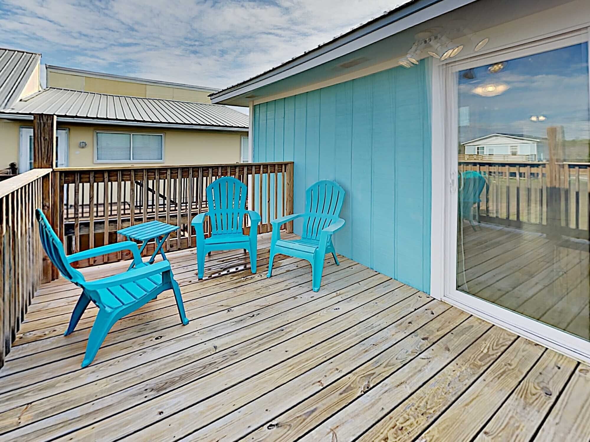 A wooden deck with turquoise chairs and a table, a sliding glass door, and a blue exterior wall.