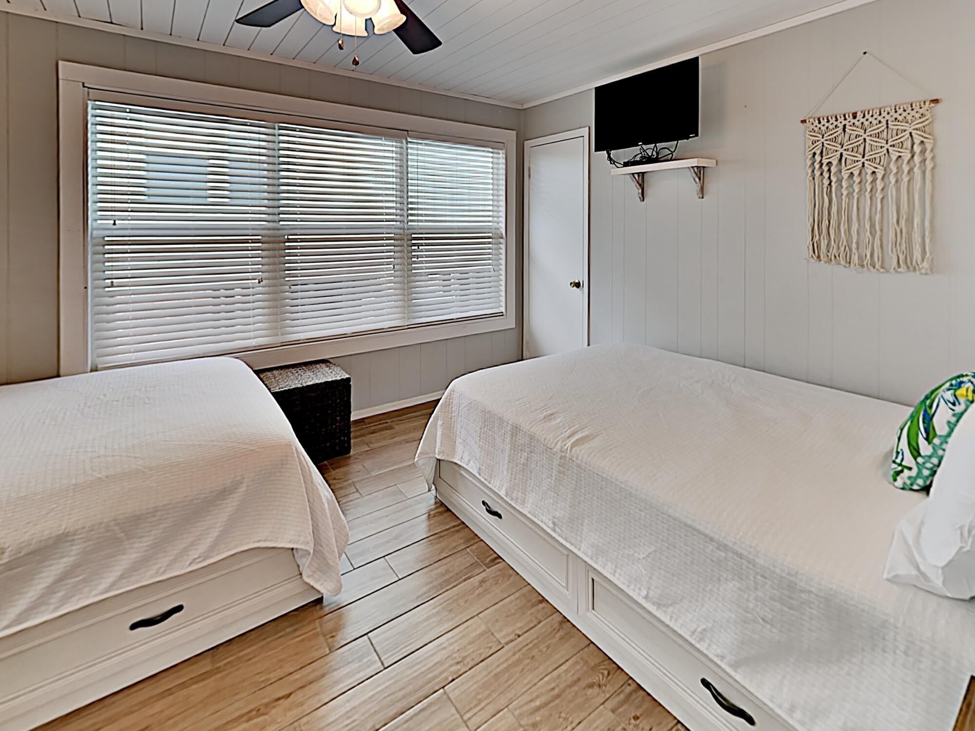 A bedroom with two beds, a ceiling fan, a TV mounted on the wall, and a macrame wall hanging.