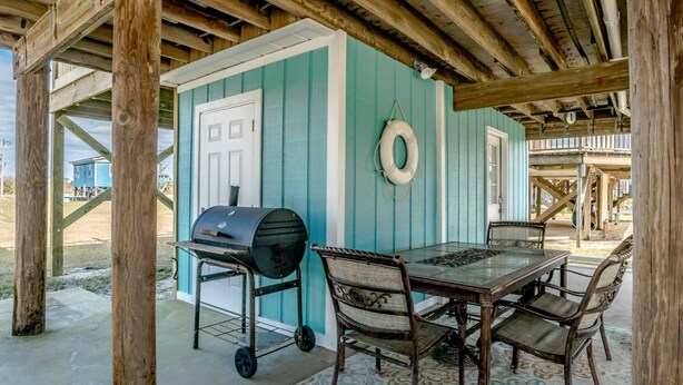 A covered outdoor area with a table and chairs, a grill, and a small building with teal walls and a white door. - Big Blue by Southern Vacation Rentals (Gulf Shores)