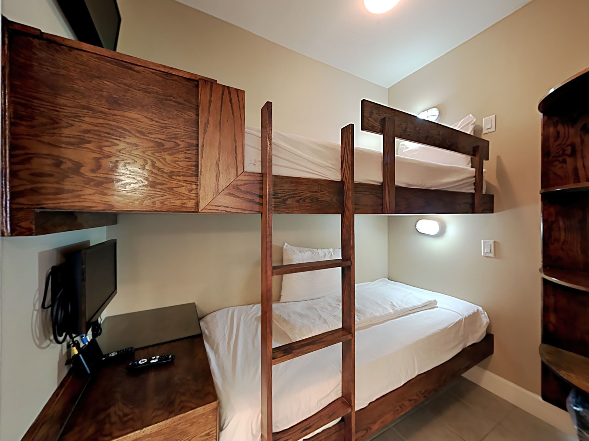 Una cama litera con escalera de madera, un escritorio con una computadora portátil y controles remotos, y un televisor instalado en la pared.