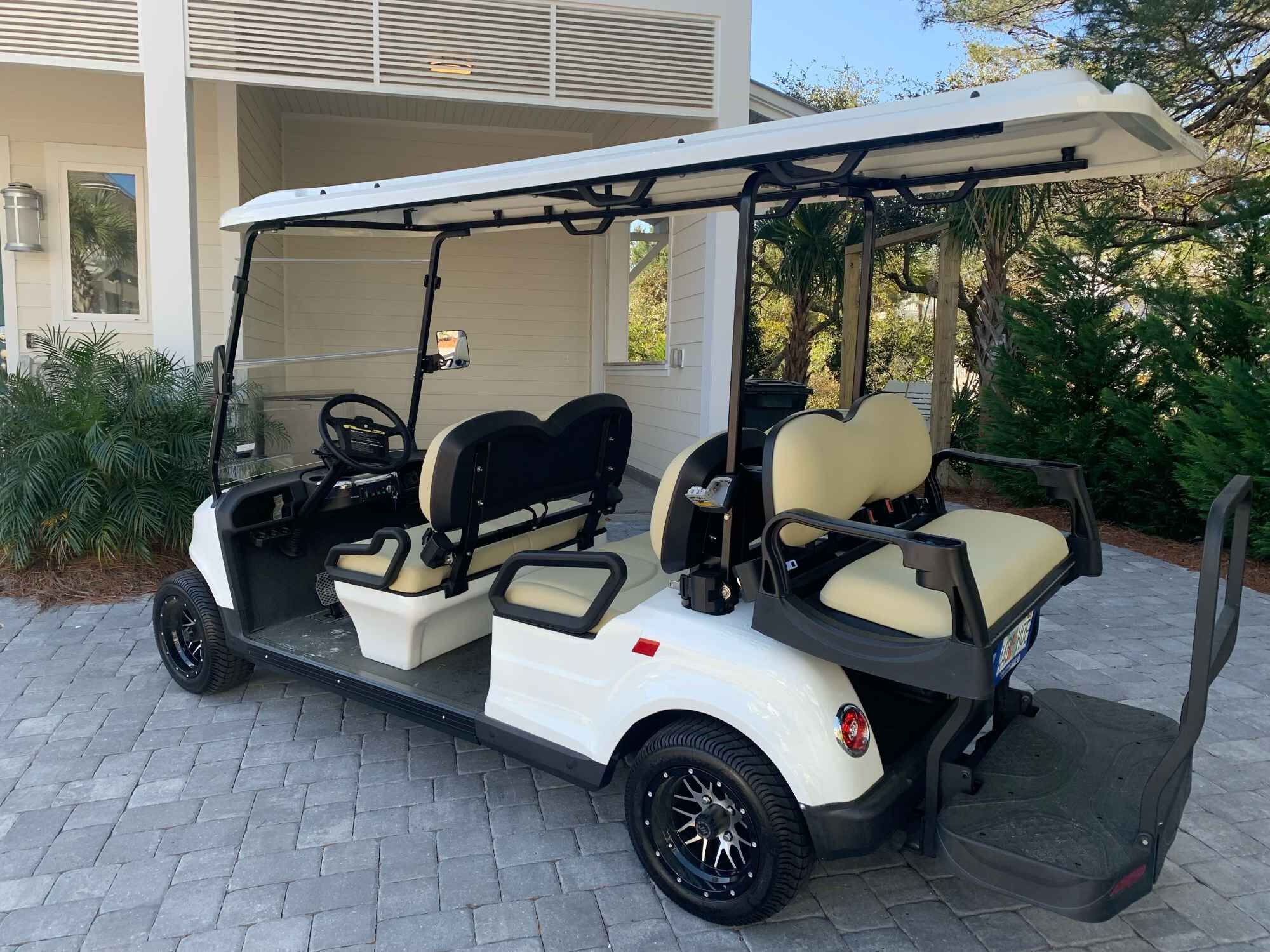 En golfbil parkerad på en asfalterad uppfart, med vit kaross och svarta detaljer, placerad framför en byggnad med en överbyggd veranda.