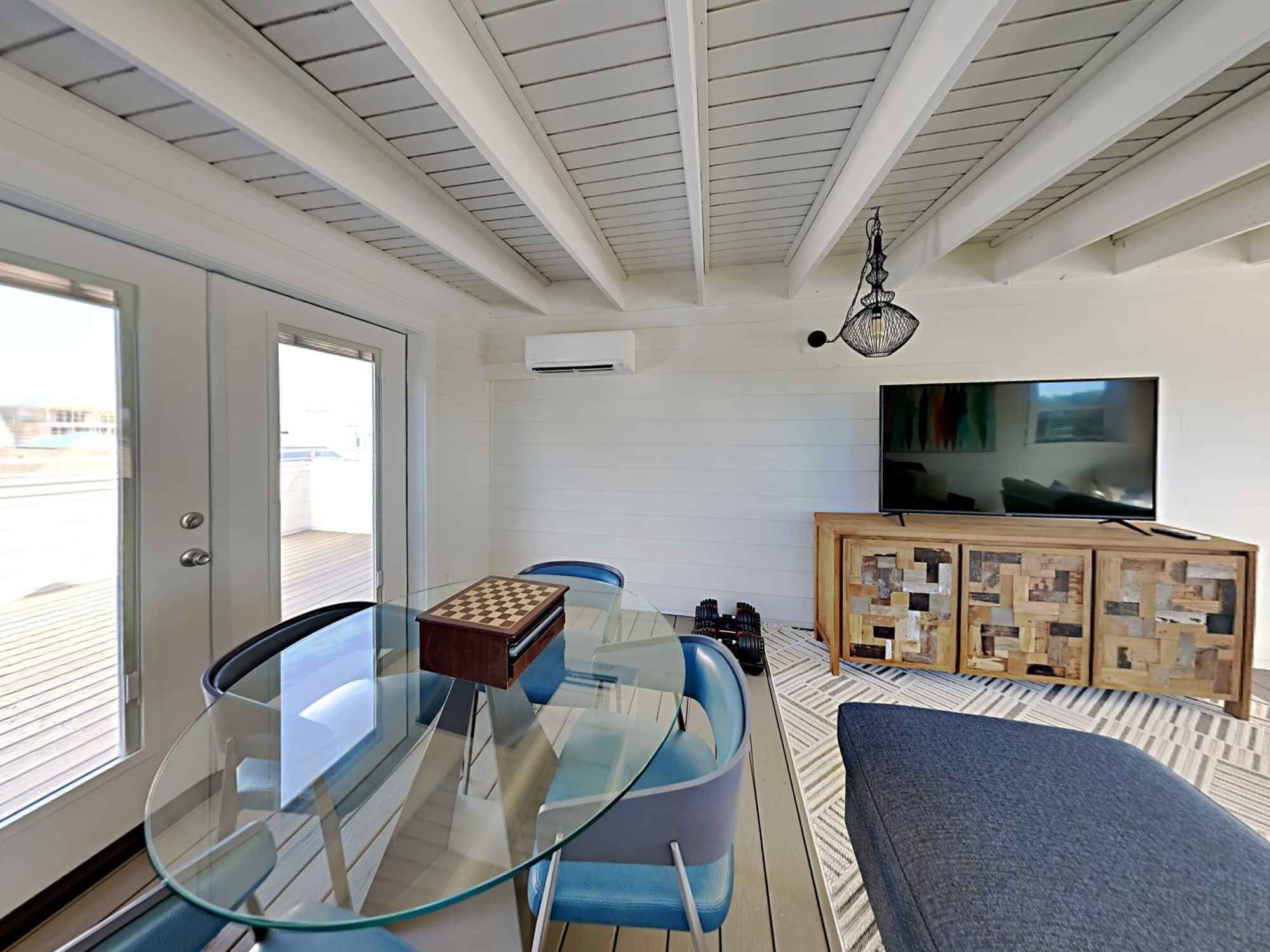 A modern living room with a glass table, blue chairs, a wooden cabinet with a TV, and a view through a glass door.