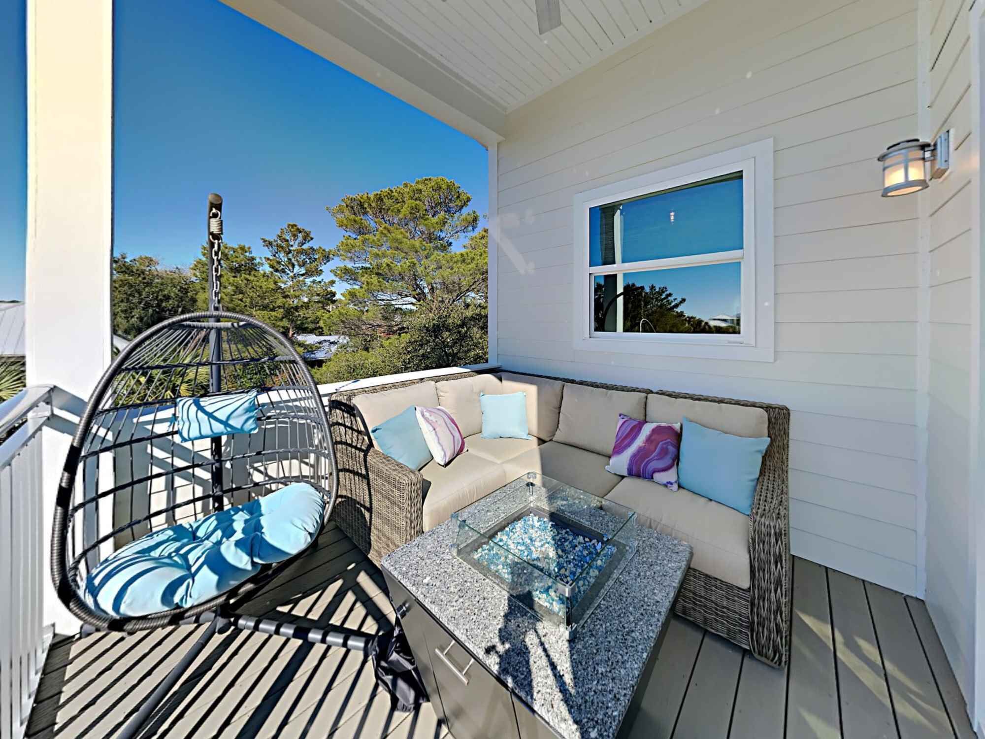 A covered outdoor seating area with a wicker chair, a glass-top table, and a sofa with cushions.