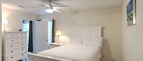 A bedroom with a white bed, a nightstand, a dresser, a ceiling fan, and a door leading to another room.