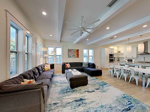 A spacious living room with a large sectional sofa, a coffee table, and a dining area with white chairs and a kitchen in the background.