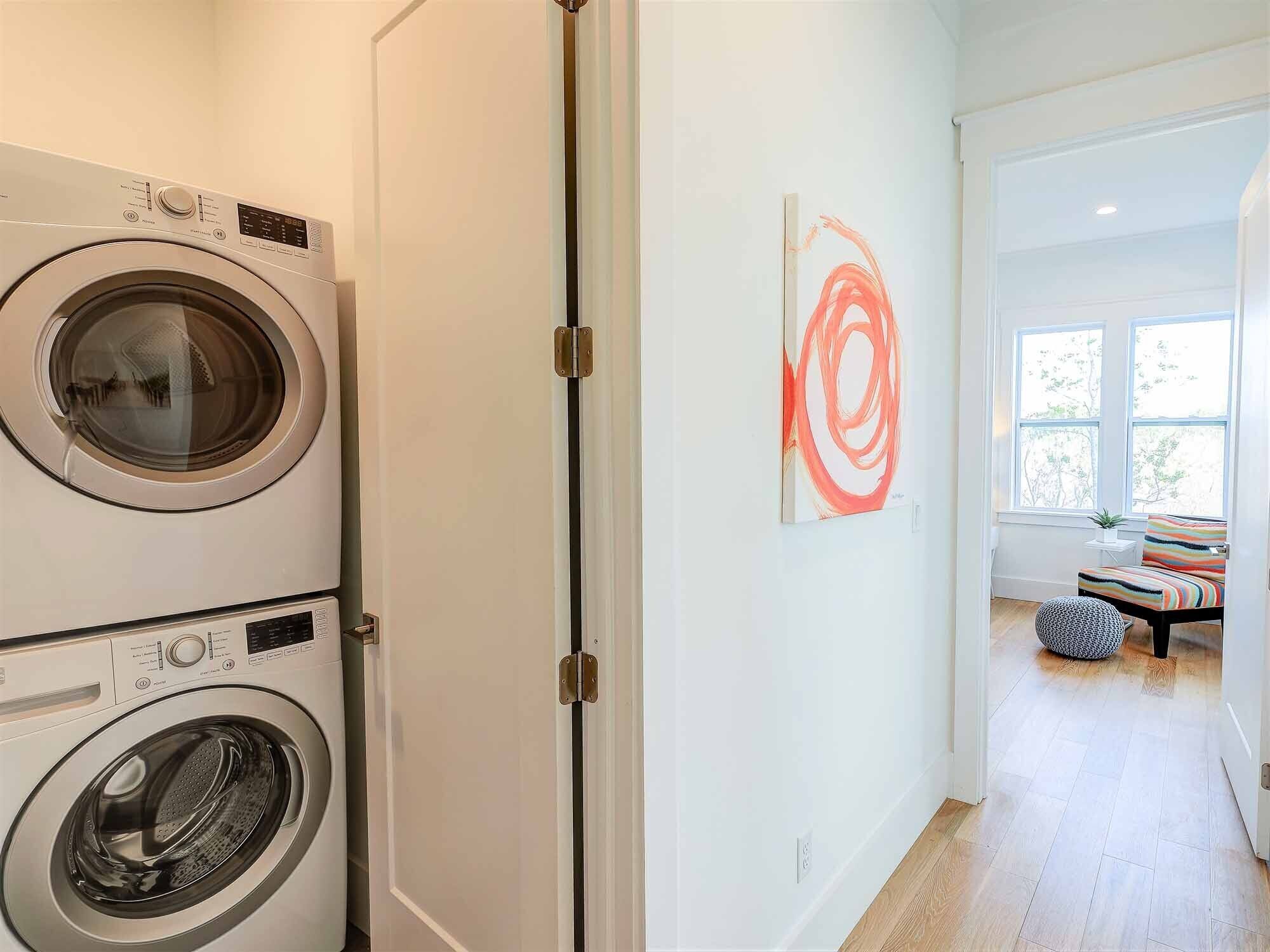A laundry room with two front-loading washing machines, a white door, and a view into a living area with a window, a chair, and a small table.