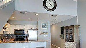 A modern kitchen with white cabinets, a stainless steel microwave, and a refrigerator. There is a clock on the wall and a bar area with stools. - Crystal Sands #311A by Southern Vacation Rentals (Destin)