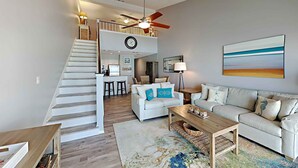 A spacious living area with a staircase leading to a second floor, a dining area, and a large painting on the wall. - Crystal Sands #311A by Southern Vacation Rentals (Destin)