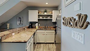 A kitchen with a granite countertop, white cabinets, a stainless steel refrigerator, and a backsplash with gray tiles. - Crystal Sands #311A by Southern Vacation Rentals (Destin)