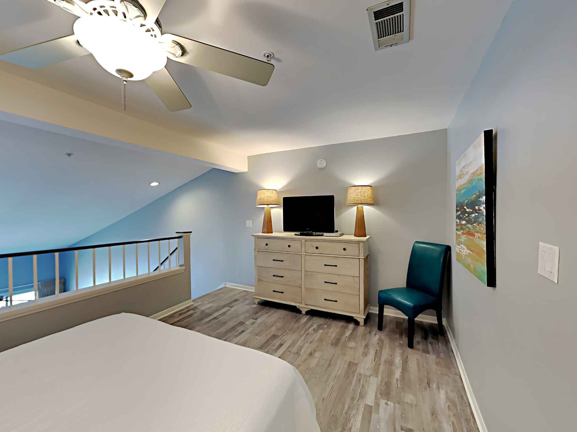 Une chambre d’hôtel avec un lit, un bureau avec une télévision, une chaise, un ventilateur de plafond et un tableau au mur.