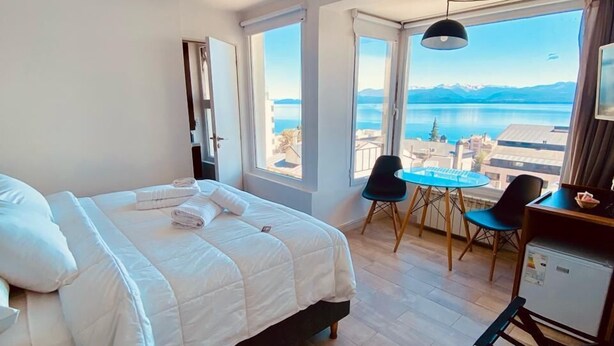A bedroom with a bed, a desk, and a chair. There are large windows with a view of a body of water and mountains. - Hotel Patagonia Signature (San Carlos de Bariloche)
