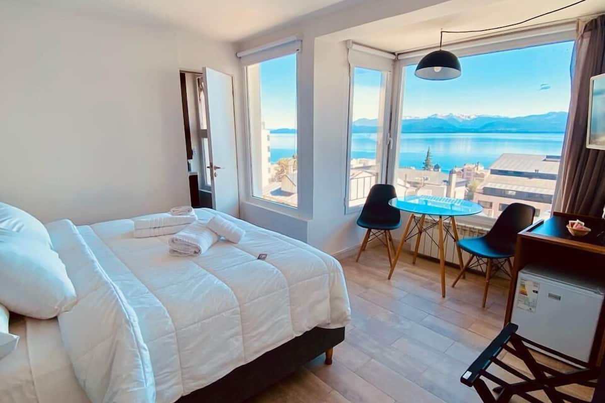 Ett sovrum med en säng, ett skrivbord och en stol. Det finns stora fönster med utsikt över en sjö eller ett hav och berg.