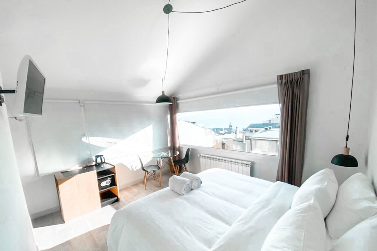 A modern hotel room with a large bed, a desk, a chair, a TV, and a view of the city.