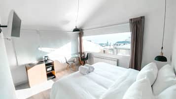 Ein modernes Hotelzimmer mit einem großen Bett, einem Schreibtisch mit Stuhl, einem kleinen Tisch und Blick auf die Stadt durch große Fenster.