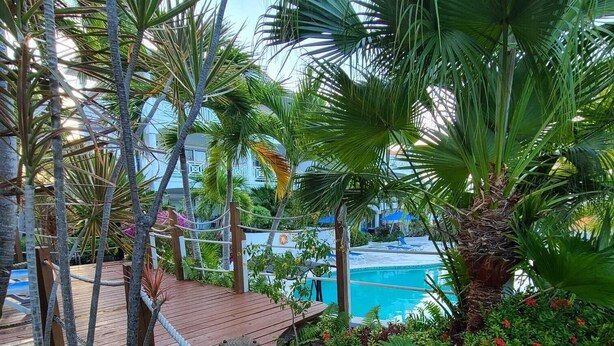 A wooden deck with a rope barrier, surrounded by lush tropical plants and a view of a swimming pool. - Queen Angel Condo with Pool and Ocean View (Providenciales)