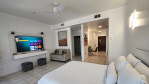A modern hotel room with a large bed, a flat-screen TV mounted on the wall, a seating area with a sofa, and a dining table. - Queen Angel Condo with Pool and Ocean View (Providenciales)