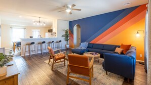 A modern living room with a blue sofa, orange armchairs, and a coffee table. There is a kitchen area with bar stools and a ceiling fan.