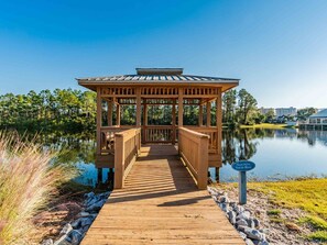 Un belvédère en bois, doté d’une allée couverte menant à un lac, est entouré d’arbres et surplombé par un ciel dégagé.