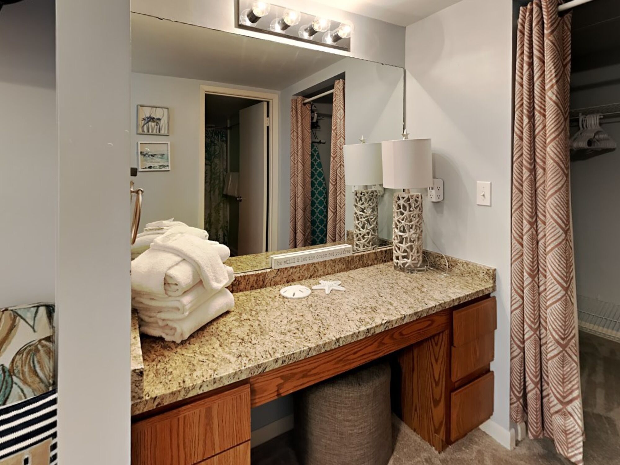 A bathroom with a granite countertop, wooden cabinets, a vanity mirror with lights, two decorative vases, and a towel stack.