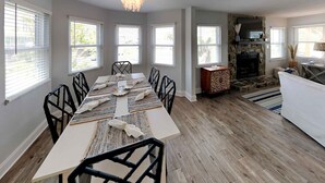 A dining area with a long table, black chairs, and a chandelier. There are windows with blinds, a fireplace, and a rug on the floor.