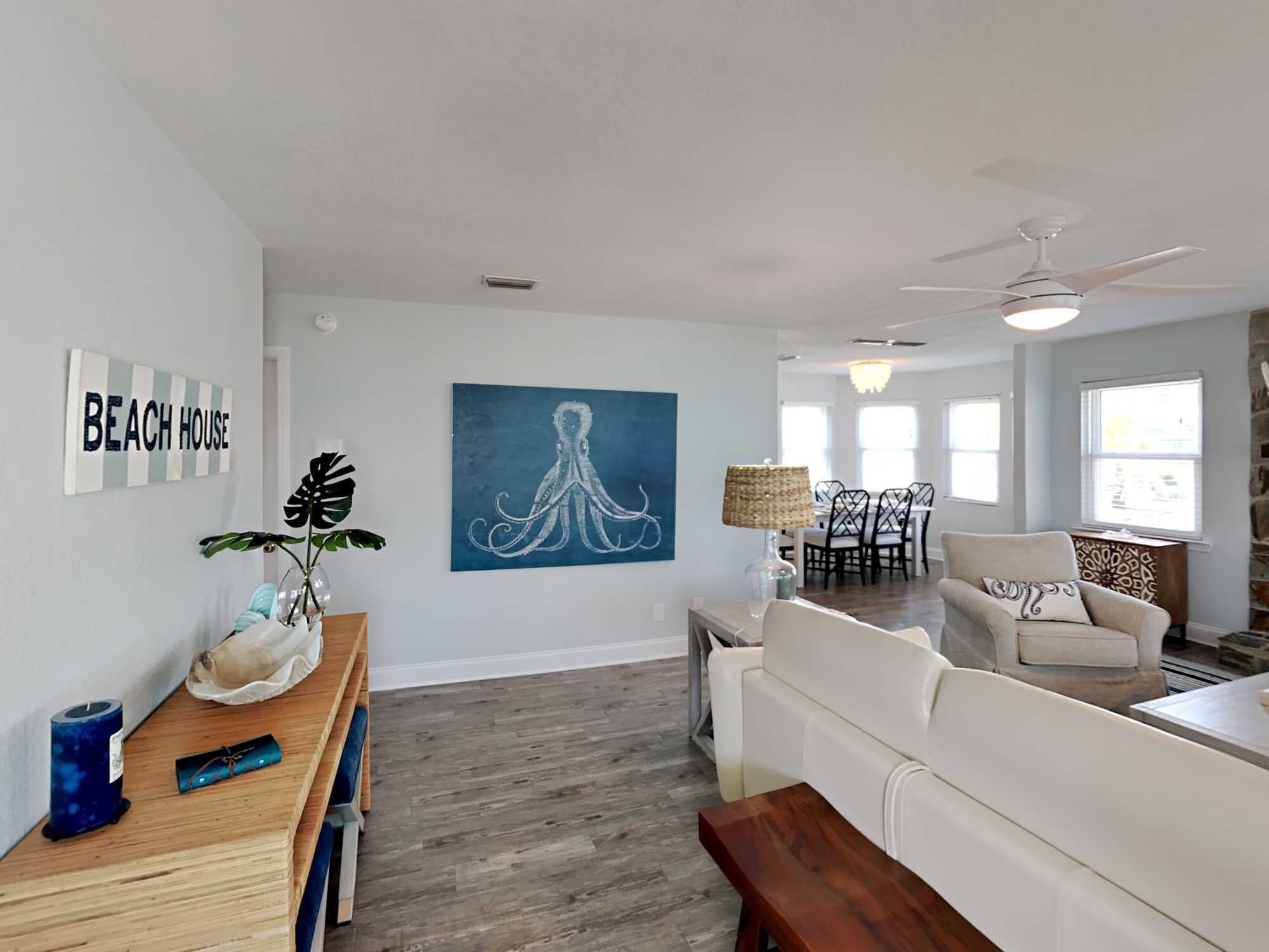 A spacious living room with a white couch, a wooden coffee table, and a dining area with a chandelier. There is a painting of an octopus on the wall and a sign that reads "BEACH HOUSE".