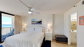 Habitación de hotel con cama, ventilador de techo, un cuadro, mesita de noche, lámpara, banco y vista a la playa.