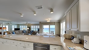 A spacious kitchen with a large island, white cabinetry, and a dishwasher. There is a dining area with a table and chairs, and a view of the ocean through large windows. - Emerald Dunes #405 by Southern Vacation Rentals (Destin)