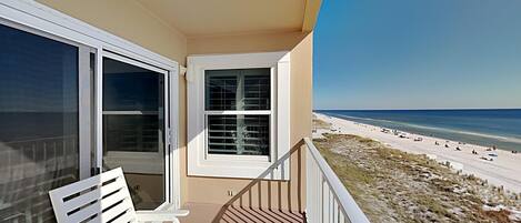 A balcony with a view of the beach, white chairs, and a window with blinds.
