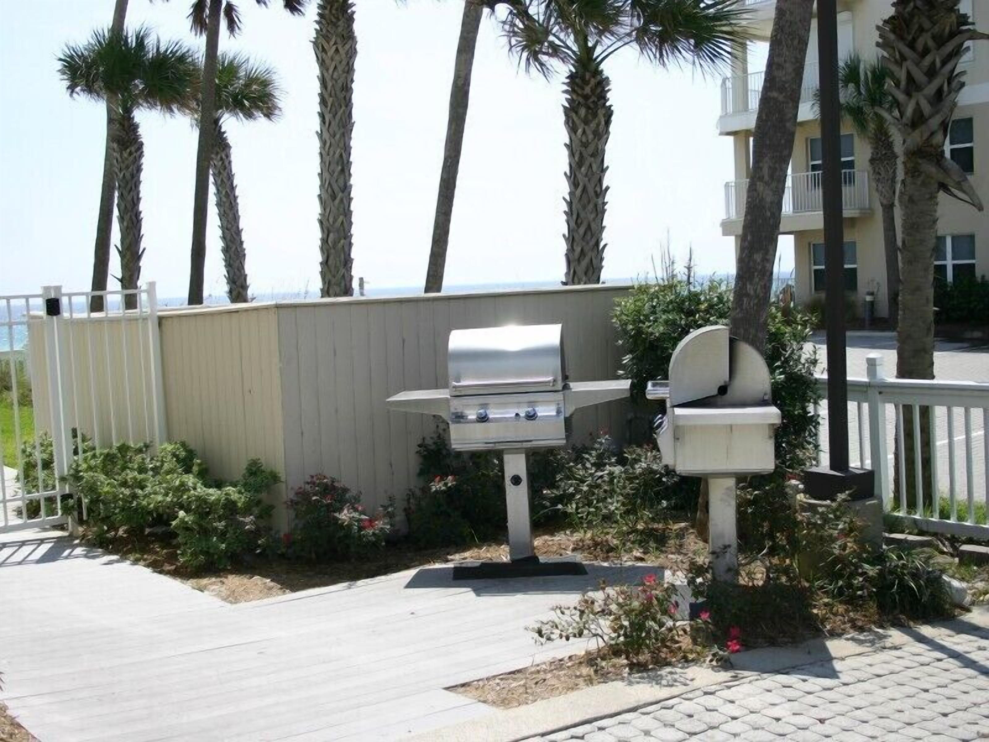 A barbecue grill and a trash bin in a landscaped area with palm trees and a view of the ocean.